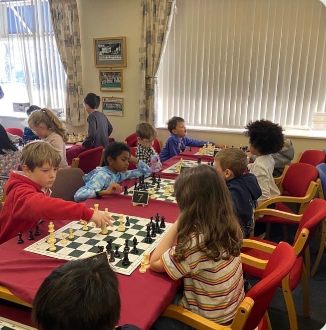 Diverse group of children playing chess at long tables in a brightly lit room.