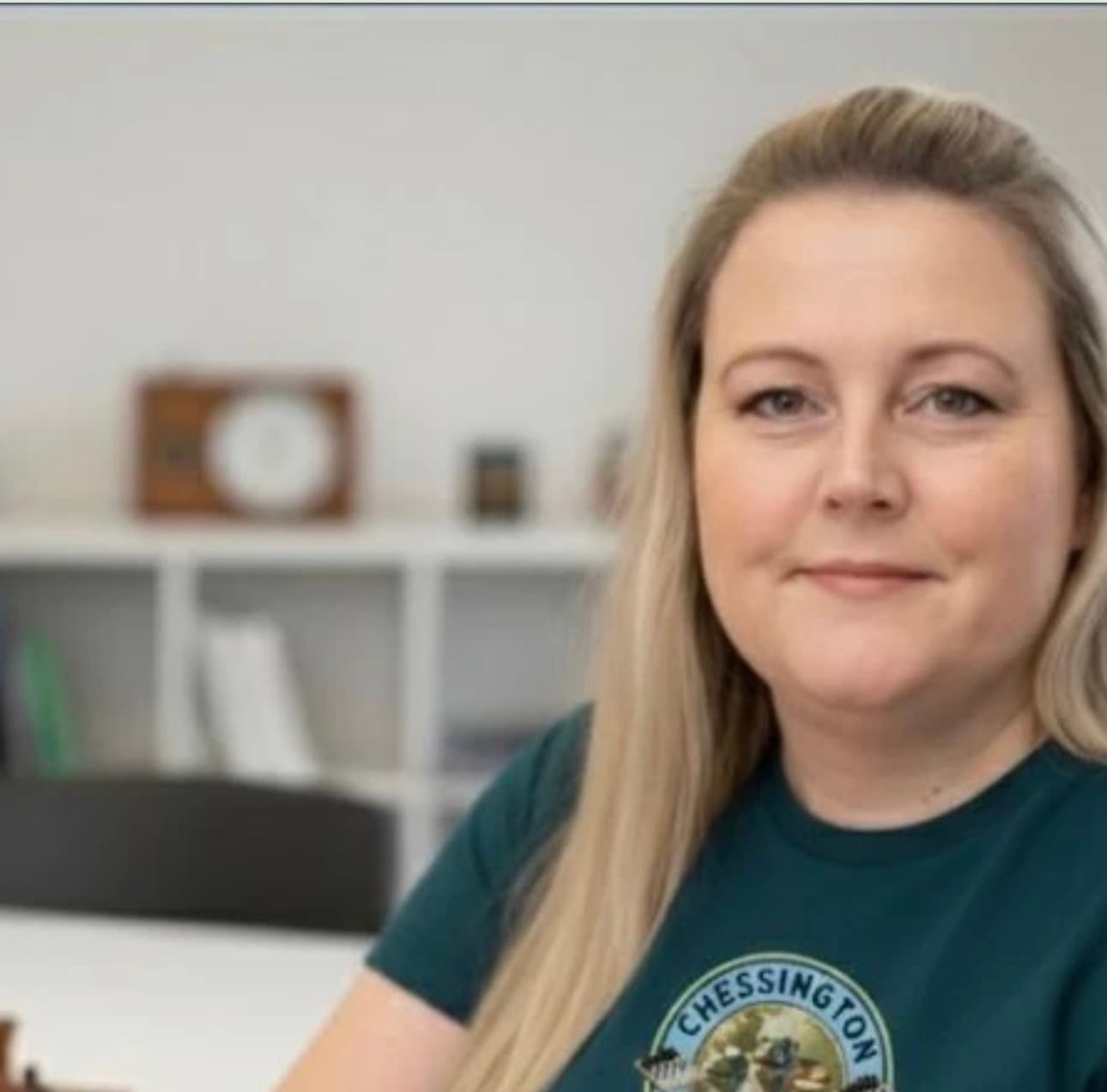 Blonde woman smiling while wearing a dark green Chessington t-shirt in a blurred office setting.