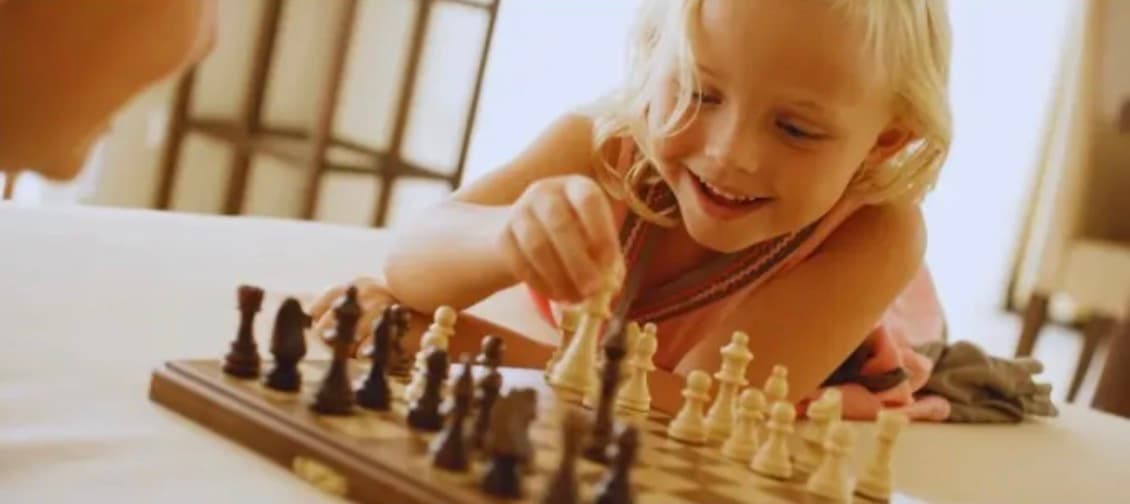 Young blonde girl smiling while moving a white piece on a wooden chess board.