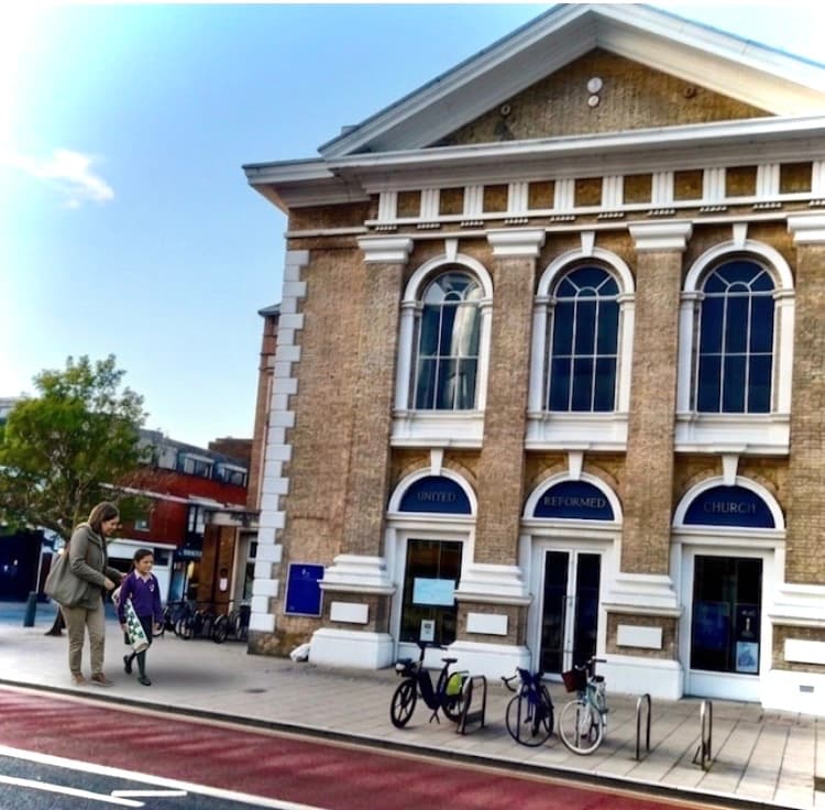Woman and child walk past a brick United Reformed Church building with arched windows.