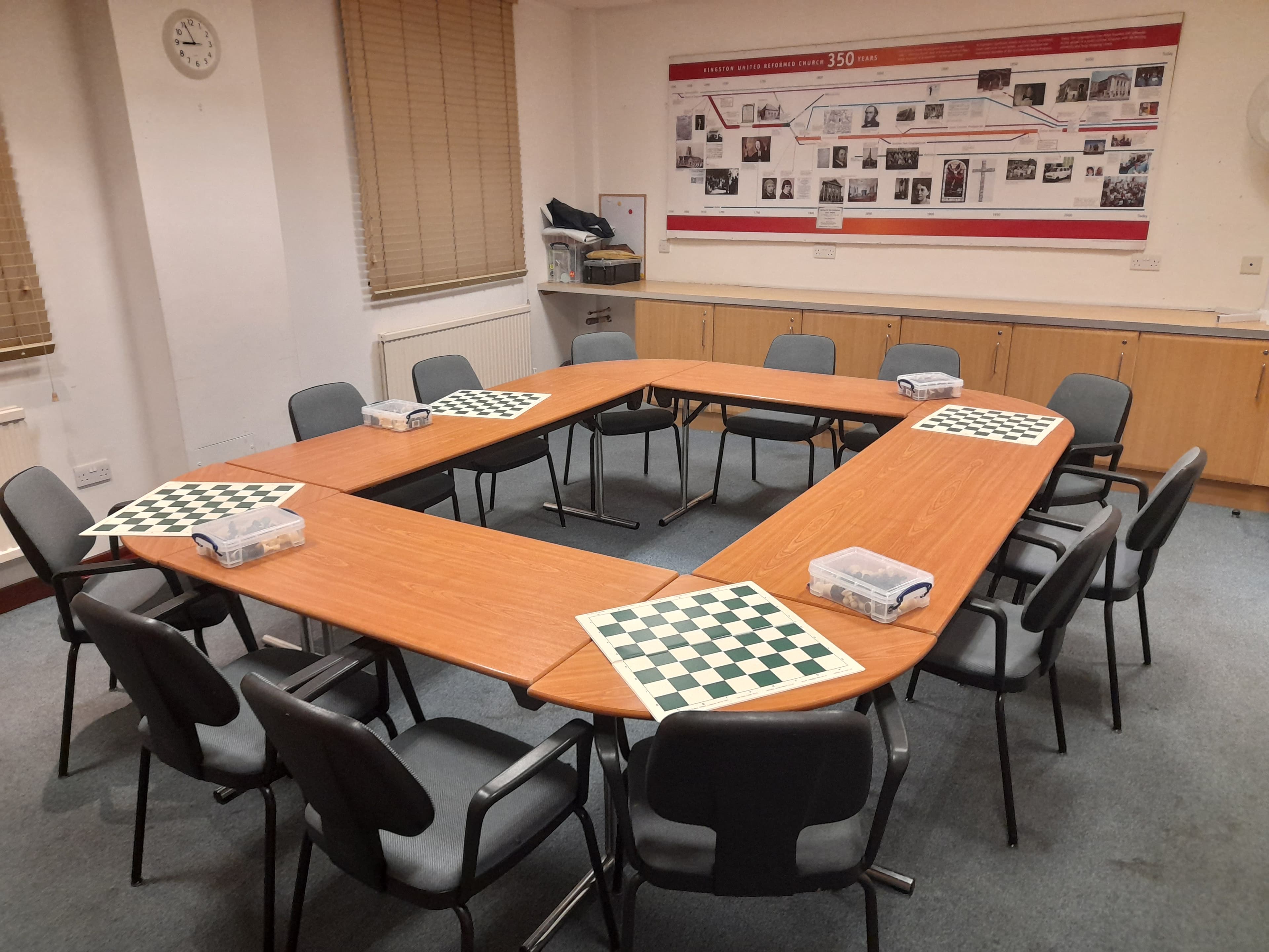 Chessboards and piece containers arranged on a U-shaped table in a room with grey chairs.