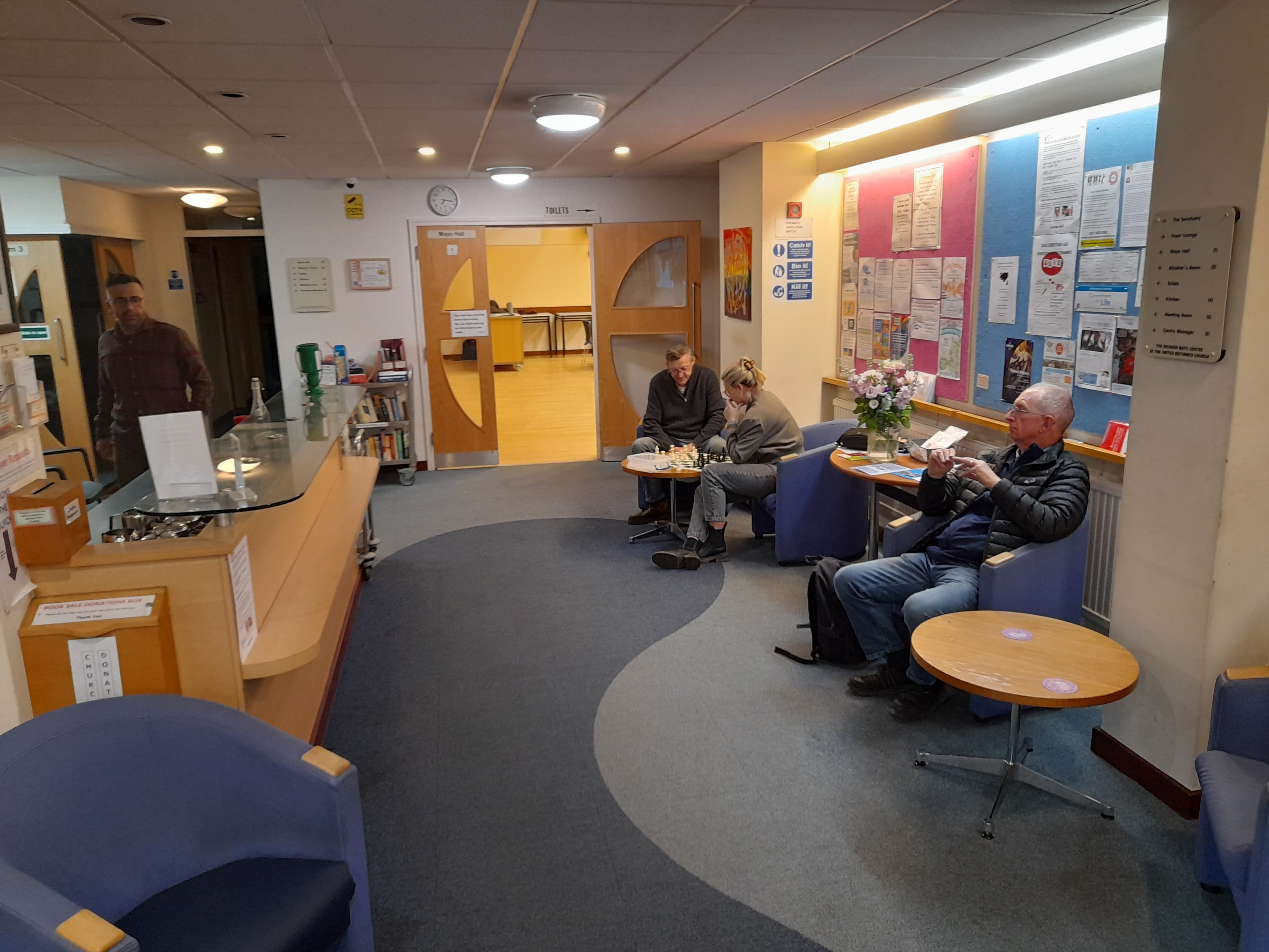 People in a community center foyer with a reception desk, notice boards, and chess players.