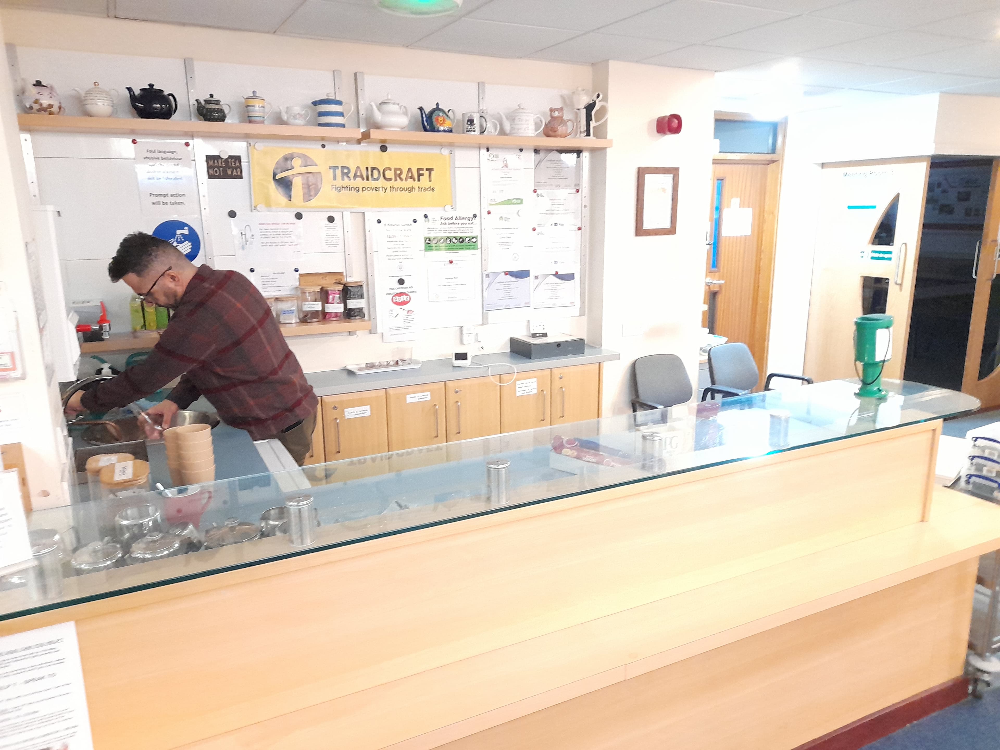 Man in a plaid shirt works behind a counter with a Traidcraft banner and teapots.